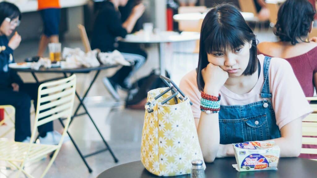 teenager who doesn't want to talk wearing striped shirt and blur denim overalls resting her head on her fist sitting at table